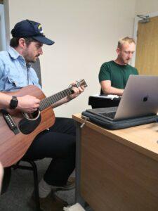 Musician James Dee is playing his guitar and to the side of him Made by Mortals Artistic Director Andy Smith plays the piano.