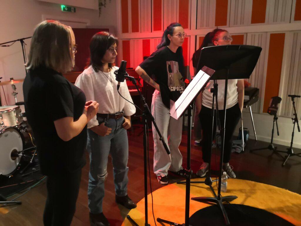 Four women standing in a recording studio with mircorphones and music stands with their scripts on in front of them.